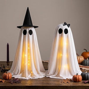 May include: Two white ghost decorations made of draped fabric, each featuring large black circular eyes. One ghost wears a black pointed witch hat, while the other has a small black bow. Both are illuminated from within by warm yellow string lights.