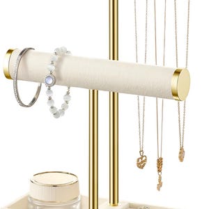 May include: A two-tier jewelry display stand with gold metal poles and beige velvet-covered bars. The stand features a rectangular base with ring slots and a small clear jar. It holds a silver bracelet, a beaded bracelet, and three gold necklaces.