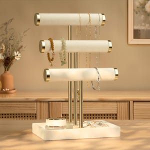 May include: A three-tier jewelry display stand featuring gold-toned metal vertical supports and horizontal bars covered in cream-colored velvet. The base is a rectangular cream-colored tray. Several necklaces and bracelets hang from the bars.