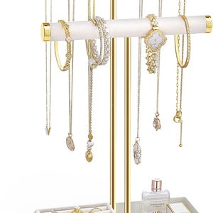 May include: A two-tier jewelry stand with gold metal poles and white velvet-covered bars. The stand holds various gold necklaces, bracelets, and a watch. A white tray at the base contains multiple rings, a perfume bottle, and a small white box.