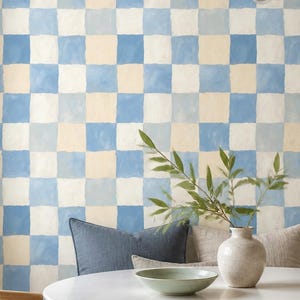May include: Wallpaper featuring a hand-painted checkerboard pattern in alternating squares of light blue and cream. The design covers the wall behind a white dining table, a wooden chair with a woven seat, and a bench with gray cushions.