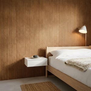 May include: A bedroom interior featuring a light wood bed frame with white bedding and a chunky knit throw, a white floating nightstand with a dark stone object, and a wall lamp. The wall is covered in vertical brown ribbed wood paneling.