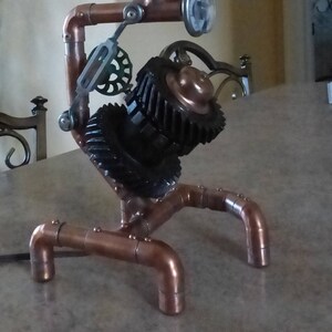 May include: A steampunk-style table lamp with a copper pipe base and a cage light fixture. The lamp features a gear mechanism and a black metal shade.