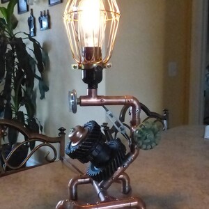 May include: A steampunk-style table lamp with a copper pipe base and a cage-style light fixture. The base features gears and other industrial-inspired elements.