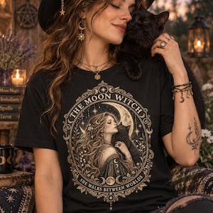 Op de afbeelding: Een zwart t-shirt met een goud- en crèmekleurige grafische print van een persoon met lang haar die een zwarte kat vasthoudt. Het ontwerp is gevat in een sierlijk ovaal kader met de tekst "THE MOON WITCH" en "SHE WALKS BETWEEN WORLDS".