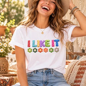 Op de afbeelding: Een wit t-shirt met korte mouwen voorzien van de tekst "I LIKE IT" in kleurrijke, gedessineerde letters in driedimensionale stijl, geplaatst boven een rij van zes gestileerde bloemvormen in diverse kleuren en patronen.