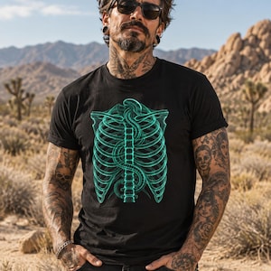 Könnte beinhalten: Ein schwarzes Kurzarm-T-Shirt mit einer leuchtend türkisfarbenen Grafik eines menschlichen Brustkorbs und einer Wirbelsäule auf der Brust. Das Design zeigt die Skelettstruktur mit einer schlangenartigen Form, die sich zwischen den Rippen windet.