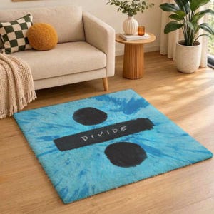May include: A rectangular area rug featuring a blue tie-dye pattern with a central black horizontal bar and two black circles. The design includes the word DIVIDE written in white capital letters across the centre of the black horizontal bar.
