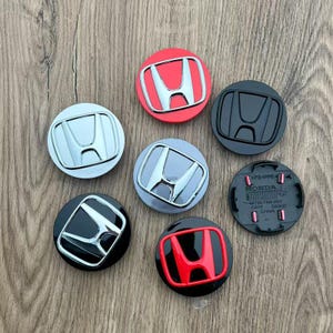 May include: Seven circular Honda wheel center caps in various finishes including chrome, matte black, red with chrome logo, and silver. One cap is flipped over to show the black plastic underside with Honda branding and part number 44732-T4N-H01.