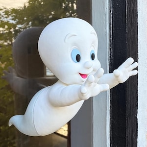 May include: A white ghost figurine with a smooth, rounded head and body, featuring large light blue eyes, arched eyebrows, and a small open mouth with a pink tongue. The figure is positioned with both arms extended forward as if reaching out.