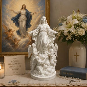 May include: A white resin statue of the Virgin Mary standing on clouds with three angels, positioned near a framed religious print, a white vase with flowers, a small sign reading My Soul Proclaims The Greatness Of The Lord, and a Holy Bible.