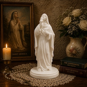 May include: A white statue of a figure in a hooded robe standing on a circular base. The figure holds a book in one hand and raises the other towards the face. The statue is positioned on a round lace doily with a string of beads nearby.