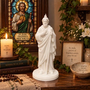 May include: A white statue of Saint Jude Thaddeus standing on a circular base, holding a staff and a medallion. The figure features a flame emblem on the head and draped robes. The statue is presented in a setting with religious decor and text.