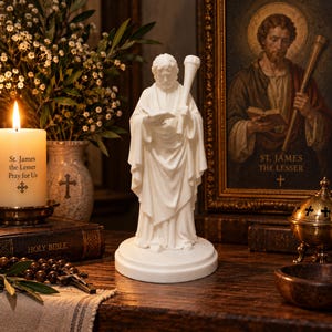 May include: A white statue of a figure holding a book and a staff, standing on a circular base. The figure wears long, draped robes. The statue is positioned in front of a framed religious portrait and a lit candle with the text St. James the Lesser.