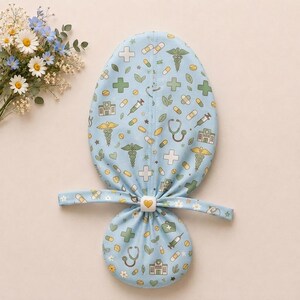May include: A light blue scrub cap featuring a repeating pattern of medical icons including stethoscopes, syringes, pills, bandages, hospitals, and caduceus symbols. The cap has a central fabric tie secured by a white button with a yellow heart.