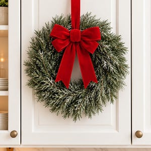 May include: A circular wreath made of dark green pine needles with a light dusting of white artificial snow. A large, vibrant red velvet ribbon is tied in a bow at the top, with two long tails hanging down over the center of the wreath.