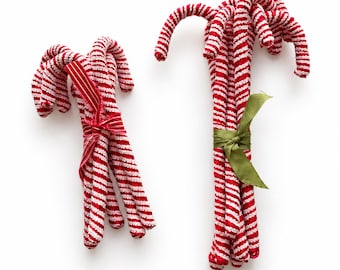 May include: Two bundles of fabric candy canes featuring a red and white striped pattern. The smaller bundle is tied with a red and white striped ribbon, while the larger bundle is secured with a solid olive green ribbon.
