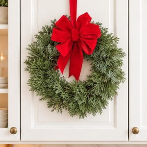 May include: A circular wreath made of dense, green, needle-like foliage, featuring a large, vibrant red velvet bow at the top. A long red velvet ribbon extends upward from the bow, allowing the wreath to hang against a white cabinet door.