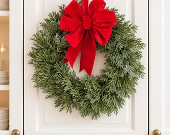 May include: A circular wreath made of dense, green, needle-like foliage, featuring a large, vibrant red velvet bow at the top. A long red velvet ribbon extends upwards from the bow, allowing the wreath to hang against a white cabinet door.