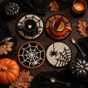 May include: Four circular crochet coasters featuring spider web and spider motifs in black, white, and orange. Two coasters support mugs, one containing a dark liquid and the other an orange liquid with a citrus slice. The set includes two spoons.