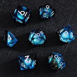 Peut inclure: Un ensemble de sept dés polyédriques présentant un intérieur translucide bleu profond et vert inspiré d'une galaxie avec des particules de paillettes en suspension. Chaque dé a des bords tranchants et des chiffres argentés bien lisibles.