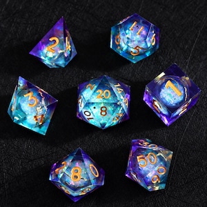 Peut inclure: Un ensemble de sept dés polyédriques en résine translucide violette et bleue avec un effet de paillettes irisé à l'intérieur. Chaque dé présente des chiffres gravés de couleur dorée, incluant un dé à vingt faces, un dé à douze faces et autres.