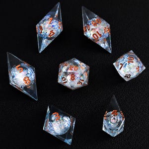 Könnte beinhalten: Ein Set aus sieben polyedrischen Würfeln aus klarem Harz mit eingebetteten irisierenden blau-weißen blumenartigen Einschlüssen. Jeder Würfel hat scharfe geometrische Kanten und kupferfarbene, eingravierte Zahlen für gute Sichtbarkeit.