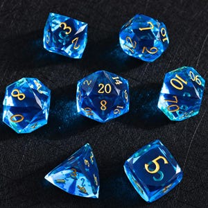 Könnte beinhalten: Ein Set aus sieben durchscheinenden blauen polyedrischen Würfeln mit goldfarbenen Zahlen. Die Sammlung enthält einen zwanzigseitigen Würfel mit der Zahl 20, einen zehnseitigen Würfel mit Zehnerzahlen, einen achtseitigen und einen zwölfseitigen Würfel.
