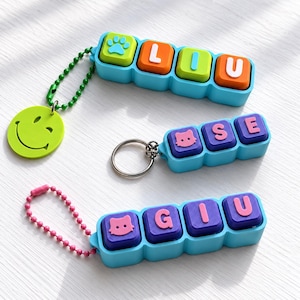 May include: Three custom keychains featuring blue plastic bases with colorful square keys. One displays a paw print and the letters LIU with a green smiley face charm. Another shows a cat face and the letters OSE. The third shows a cat face and GIU.