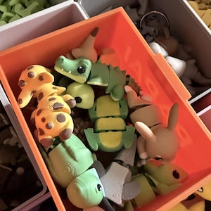 May include: An orange plastic bin filled with various small, articulated 3D-printed animal figures, including a yellow and brown giraffe, a green crocodile, a light brown rabbit, and a green bird, all featuring visible layered textures.