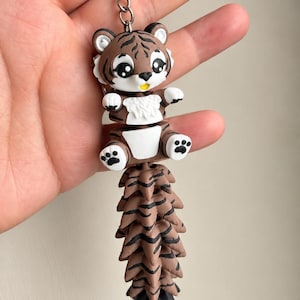 Op de afbeelding: Een sleutelhanger met een bruin-witte tijgerfiguur met grote zwarte ogen en een kleine gele mond. De tijger heeft een gesegmenteerde, beweegbare staart met bruine en zwarte strepen die aan een zilveren metalen ring en ketting is bevestigd.