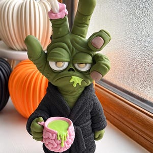 Op de afbeelding: Een groen zombieachtig figuurtje met een handvormig hoofd dat een vredesteken maakt, gekleed in een zwarte badjas met textuur. Het figuurtje heeft hangende ogen, een groene substantie die uit de mond druipt en houdt een roze hersenvormige mok vast.