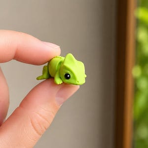 Op de afbeelding: Een miniatuur kameleonfiguurtje in limoengroen met een gladde matte afwerking, voorzien van een prominente kopkam, kleine getextureerde ledematen en één groot zwart oog. Het kleine hagedisje wordt tussen twee vingers vastgehouden.