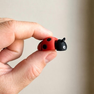 Op de afbeelding: Een klein lieveheersbeestje-figuurtje met een felrode rug met duidelijke zwarte stippen en een effen zwarte kop met twee kleine voelsprieten en één zichtbaar wit oog met een zwarte pupil, afgewerkt met een gladde, matte textuur.