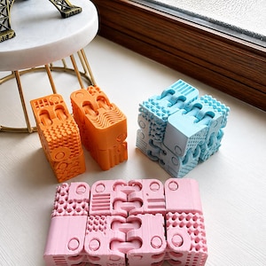 May include: Two orange, one light blue, and one light pink articulated fidget cubes. Each cube is composed of multiple interconnected segments featuring various geometric textures, including grid patterns, wavy lines, and circular indentations.