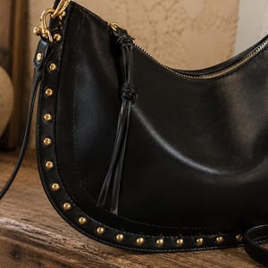 Peut inclure: Un sac à bandoulière noir en forme de croissant avec un bord inférieur incurvé orné d'une rangée de petits clous métalliques dorés. Le sac comprend une fermeture éclair dorée, une tirette en cuir noir assortie avec un nœud et une fine bandoulière.