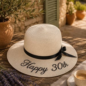 Könnte beinhalten: Ein cremefarbener Sonnenhut aus Stroh mit einem schwarzen Schleifenband und dem in Schwarz gestickten Text "Happy 30th" auf der Krempe. Der Hut hat eine abgerundete Krone und eine breite, flache Krempe für eine strukturierte Optik.
