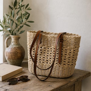 Peut inclure: Un sac cabas en paille tressée de couleur beige naturel, doté d'un motif ajouré en treillis et de deux longues anses en cuir marron. Le sac présente une base rectangulaire structurée et une doublure en tissu clair visible à l'ouverture.