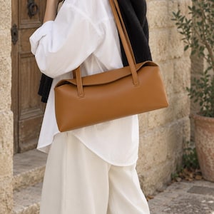 May include: A tan, rectangular shoulder bag with a smooth finish, featuring a structured horizontal silhouette, a top flap closure, and two thin, matching shoulder straps attached to the top edge of the bag for carrying over the shoulder.