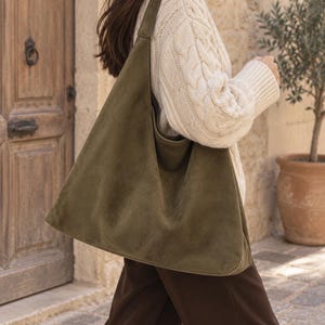 Può includere: Una grande borsa a spalla stile hobo color verde oliva con una consistenza morbida e opaca. La borsa presenta un'ampia tracolla integrata e una silhouette spaziosa e destrutturata che pende verticalmente lungo il fianco.