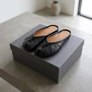 Könnte beinhalten: Ein Paar schwarze Schlupfschuhe aus Leder mit geteilter Zehenpartie, die ein kleines Knotendetail in der Mitte des Vorderblatts und ein beiges Innenfutter aufweisen und auf einem dunkelgrauen, rechteckigen Karton mit geprägtem Logo stehen.