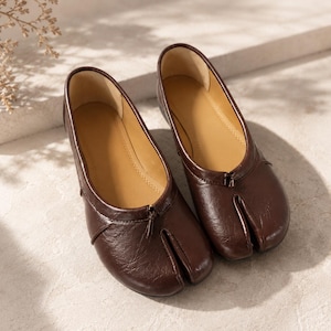 May include: A pair of dark brown leather tabi shoes featuring a split-toe design, a smooth finish, and a small decorative knot detail at the center of the vamp. The interior is lined with a tan material, and the shoes have a low-profile silhouette.