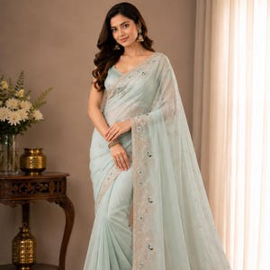 Puede incluir: Un sari de color verde menta claro confeccionado en una tela ligera y transparente con bordados intrincados a lo largo de los bordes. La prenda incluye delicados patrones florales y sutiles adornos de lentejuelas, junto a una blusa sin mangas.