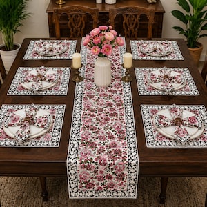 May include: A dining table set featuring a long rectangular table runner and six matching square placemats, all decorated with a pink and green floral pattern on a white background, accompanied by six white plates and six floral napkins.