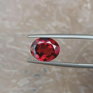 Könnte beinhalten: Ein runder, facettierter, tiefroter Edelstein, der von einer silberfarbenen Metallpinzette gehalten wird. Der Stein weist einen Brillantschliff mit zahlreichen dreieckigen und rautenförmigen Facetten auf, die das Licht reflektieren.