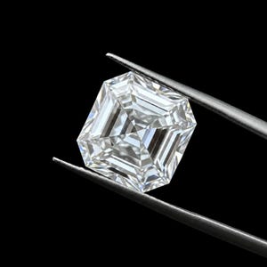 May include: A clear, colorless, square-cut gemstone with a faceted surface that reflects light, held by a pair of metal tweezers against a solid black background. The stone features a geometric, step-cut pattern with sharp, symmetrical edges.