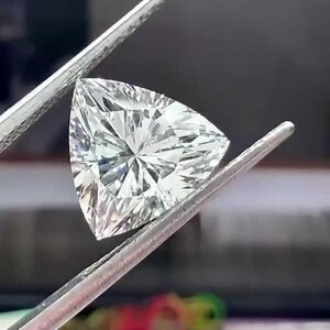 Könnte beinhalten: Ein klarer, farbloser Edelstein im Trillion-Schliff, der von einer Metallpinzette gehalten wird. Der Stein hat eine dreieckige Form mit spitzen Ecken und zahlreichen Facetten, die das Licht reflektieren und eine brillante, funkelnde Oberfläche erzeugen.