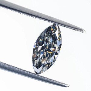 Può includere: Una pietra preziosa taglio marquise con superficie sfaccettata, che mostra una tavolozza di colori grigi e argento con elevata chiarezza e riflessione della luce, tenuta saldamente tra le due punte metalliche di una pinzetta di precisione.
