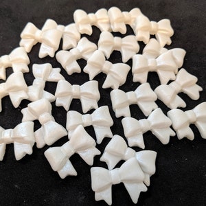 May include: A collection of white, pearl-like, sugar bow decorations. The bows are scattered on a black surface.