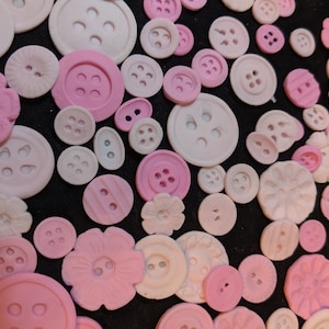 May include: A collection of pink and white buttons of various sizes and designs. The buttons are made of a smooth, shiny material and have a variety of textures, including smooth, textured, and embossed.